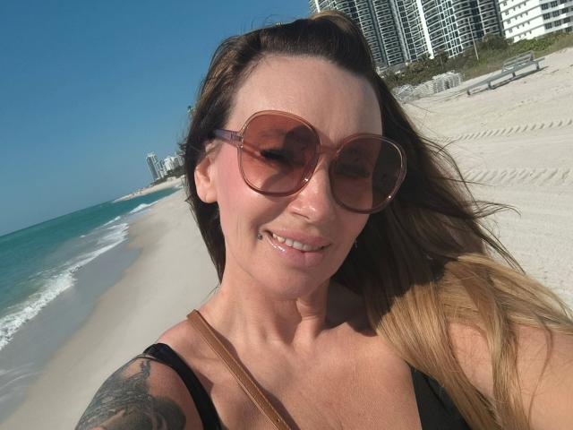 MissJenn-hot with brown sunglasses, smiling, wearing a black top, showing a tattoo on her right arm, standing on a sunny beach with blue ocean and white sand.