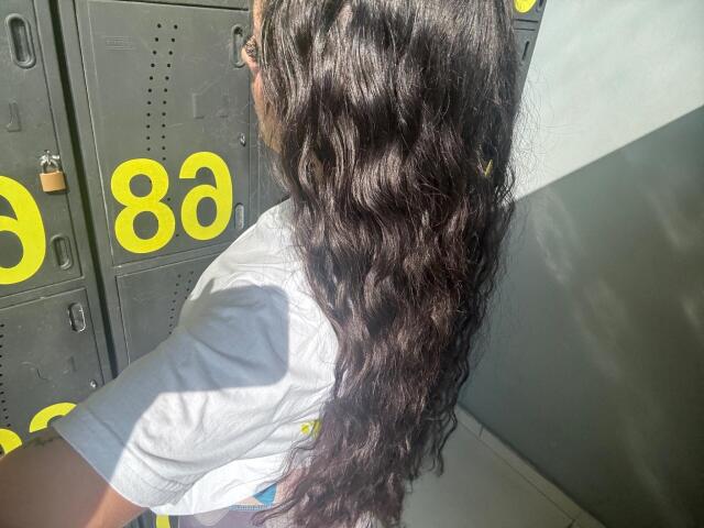 AlisonTatcher's long, wavy brown hair is in sunlight, flowing over her white shirted back, standing in front of gray metal lockers with yellow numbers.