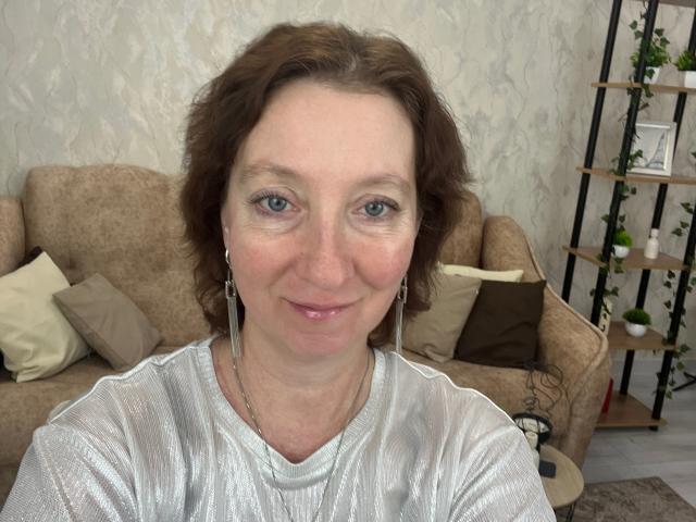 MadaamTang has short brown hair, blue eyes, and a slight smile. She's wearing a shiny silver top with earrings, and has a slightly wrinkled face. Boobs are not visible.