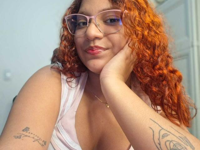 VeronicaBellerose with curly red hair, pink glasses, and a pink tank top, resting her cheek on her hand, showing tattoos on her arms, and a subtle smile.