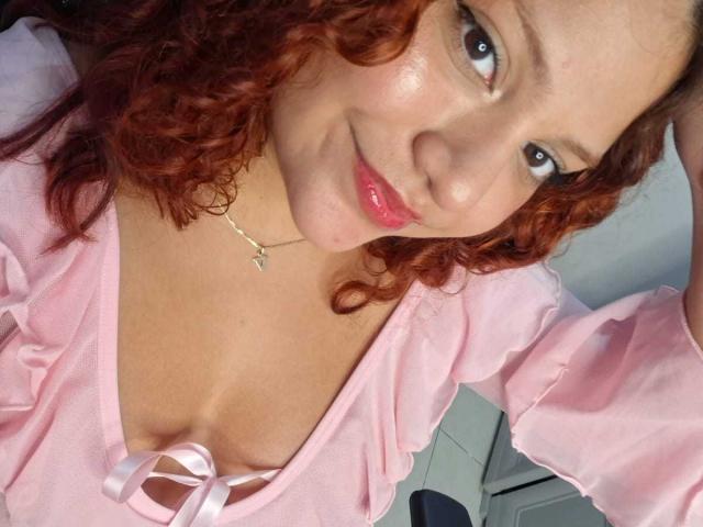 VeronicaBellerose has curly red hair, pink lips, and a light skin tone. She's wearing a pink top with a ribbon, showing her tits. She's smiling at the camera.