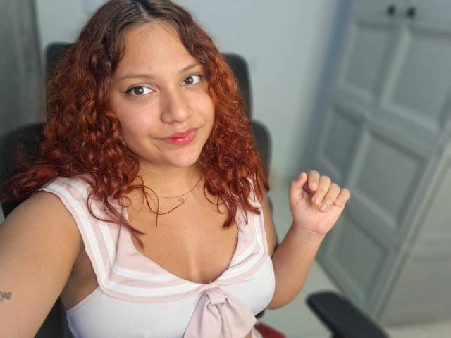 VeronicaBellerose with curly red hair, light skin, and pink lipstick, wearing a white sailor top with a pink bow, smiling, holding up a hand, showing small tits.