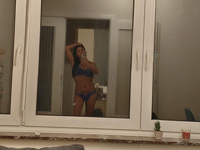 CharlotteGossart in blue lingerie, standing in front of a mirror, holding her phone with one hand, showing her tits and ass, in a dimly lit room.