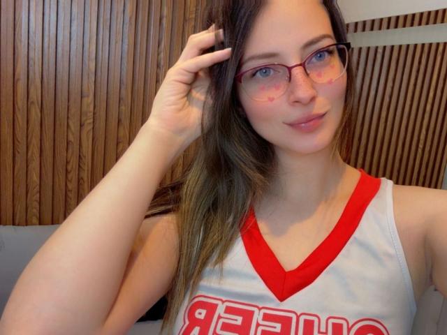AmbeerWood with glasses, wearing a red V-neck cheerleader top, showing her tits, has long brown hair, fair skin, and a slight smile, touching her hair.