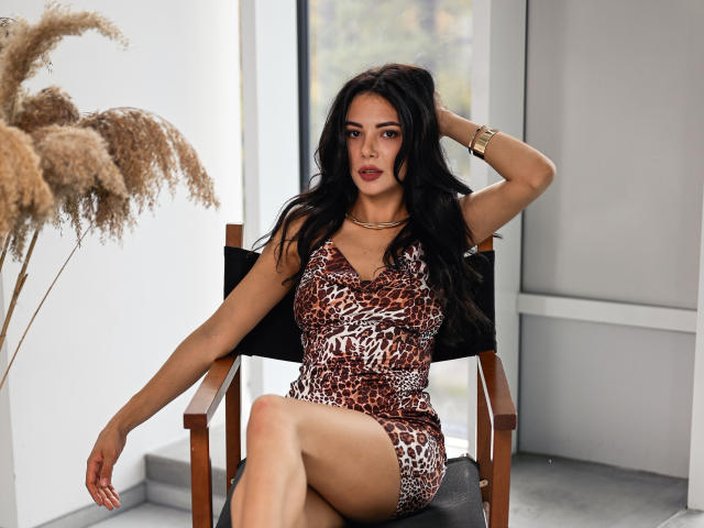 OnlyXandra with long black hair, wearing a leopard print dress, sitting on a black chair, hand in hair, gold bracelet, legs crossed, looking at camera, pouty lips.