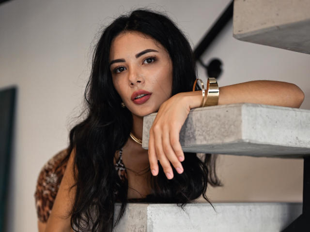 OnlyXandra with long black hair, brown eyes, and red lips, leaning on concrete stairs, wearing a gold bracelet and leopard print top, looking directly at the camera.