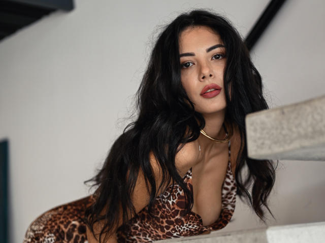 OnlyXandra mit langem, schwarzem Haar, einem Leopardenmuster-Bikini und einer goldenen Halskette, lehnt sich an eine Betontreppe, blickt mit roten Lippen und leicht geöffnetem Mund in die Kamera..