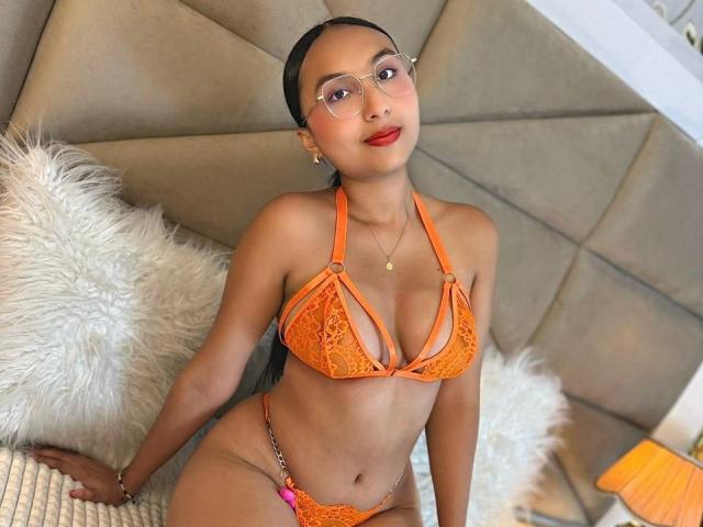 ErikaMartini wearing an orange lace bikini, showing her small tits and pussy, with a red lip and glasses, sitting on a white furry pillow.