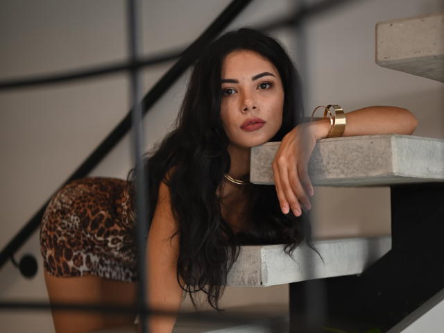 OnlyXandra mit langen, schwarzen Haaren, die leopardenmusterige kurze Hose trägt, lehnt sich an eine Betontreppe, trägt ein goldenes Armband, hat einen ernsten Gesichtsausdruck, dunkle Augenbrauen, volle Lippen und braune Augen und blickt in die Kamera..