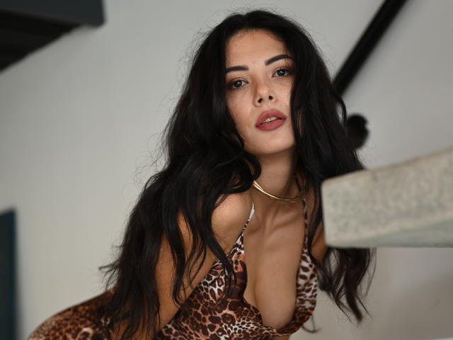 OnlyXandra with long black hair, wearing a leopard print dress, gold necklace, and red lipstick, leaning forward, showing her cleavage and looking at the camera.