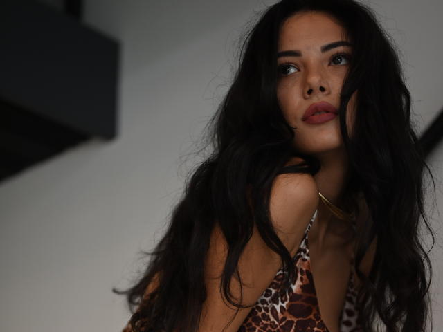 Solo Xandra, con su largo cabello negro y ondulado, vistiendo una blusa de estampado de leopardo, tiene los labios rojos y una expresión ligeramente fruncida, mirando hacia arriba con ojos oscuros y seductores. Sus senos son parcialmente visibles..