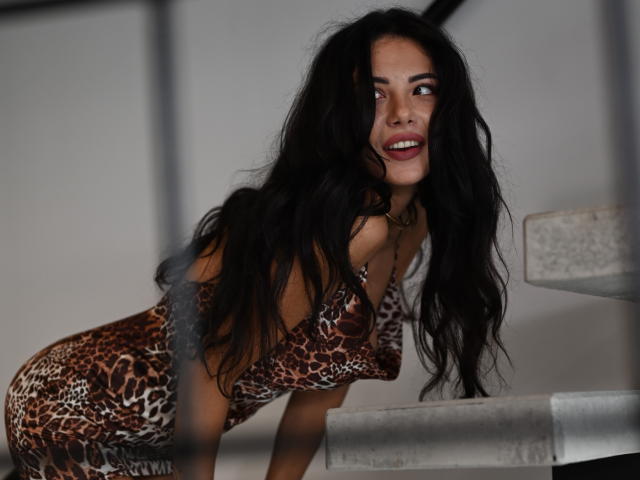 OnlyXandra with long black hair, wearing a leopard-print dress, leaning forward with a playful smile, showing her ass. She's standing next to a concrete staircase.