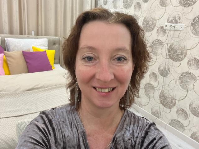MadaamTang with short brown hair, smiling, wearing a gray velvet top, showing her teeth, and earrings, sitting in a room with a bed and patterned wallpaper.