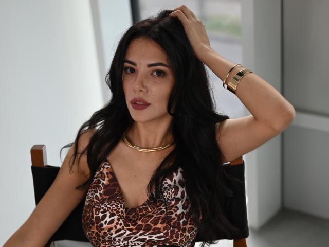 OnlyXandra met lang zwart haar, gekleed in een jurk met luipaardprint, een gouden ketting en armband, met haar hand in haar haar, zittend op een zwarte stoel, kijkend naar de camera, met een kleine neus en volle lippen..