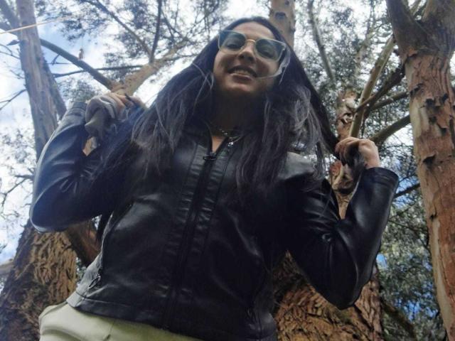 JessyLuxs with long black hair, wearing a black leather jacket, sunglasses, and green pants, standing under tall trees, smiling, and holding her hair with both hands.