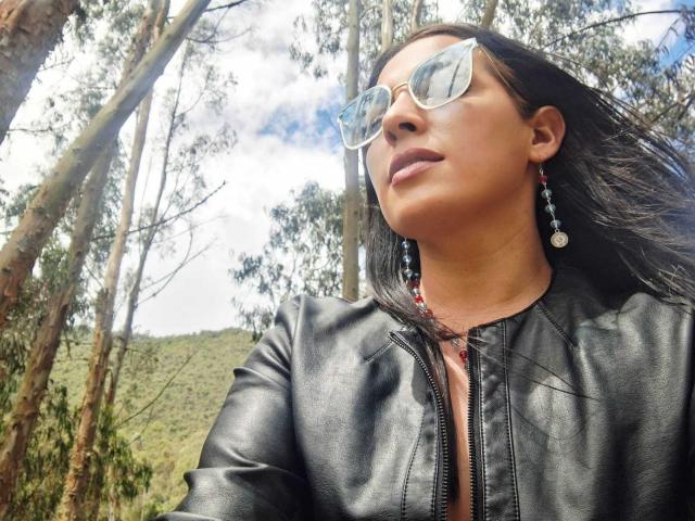 JessyLuxs with dark hair, wearing clear sunglasses, black leather jacket, and red earrings, looking up at the sky, standing in a forest, sunny day, face slightly tilted.