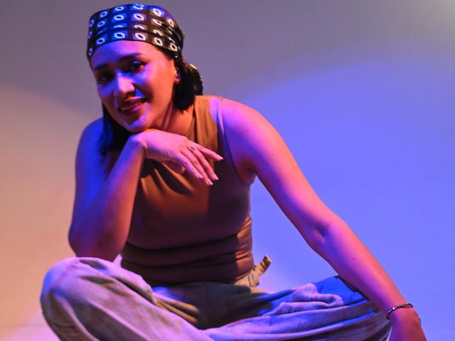 ScarlethFely with black bandana, brown tank top, and gray pants, squatting, smiling, left hand on chin, right arm on knee, blue and orange lighting, relaxed.