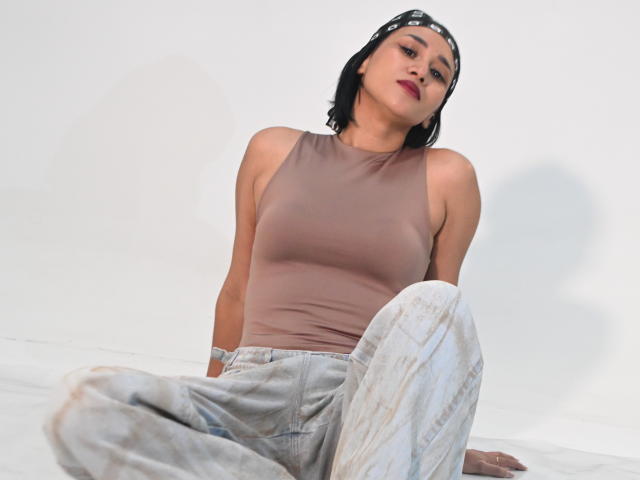 ScarlethFely with short black hair, wearing a beige tank top and dirty jeans, sitting with legs crossed, head tilted, and bandana, showing her small boobs and confident expression.