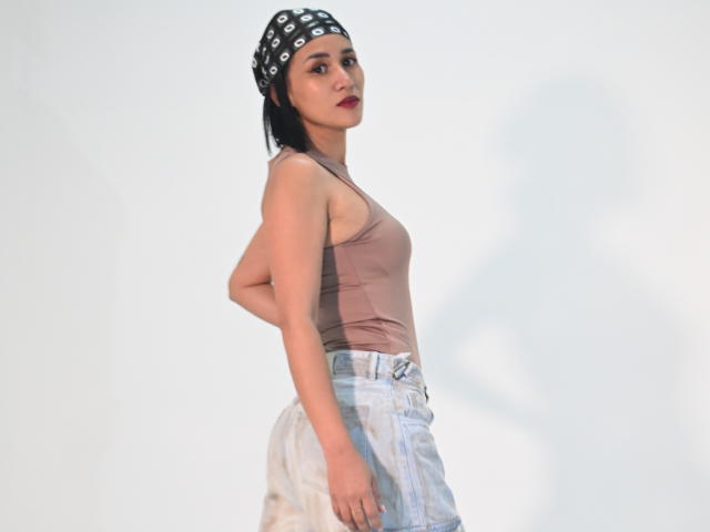 ScarlethFely has a brown tank top, white high-waisted jeans, and a black polka-dot bandana, with red lipstick and dark eyes, standing side-on with hands on her ass.