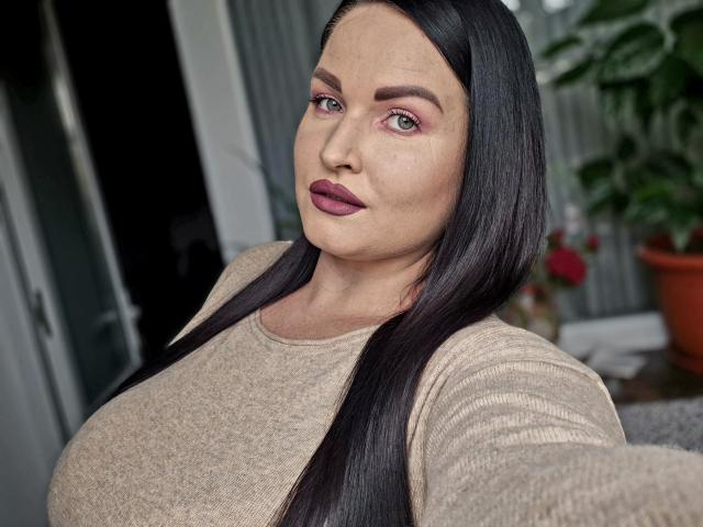 GeorgiaLerox has large boobs, long black hair, pink eyeshadow, and purple lipstick. She's wearing a beige sweater, looking at the camera with a slight smile.