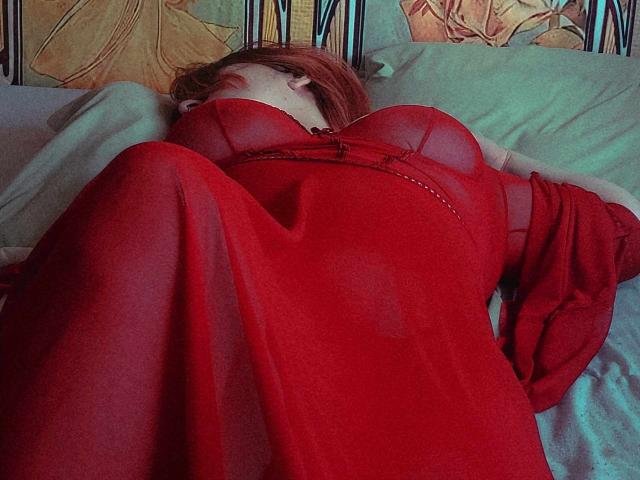 CallMeElysia lying on a bed, wearing a red sheer dress that shows her tits, with her legs up and head tilted back, red hair covering her face.
