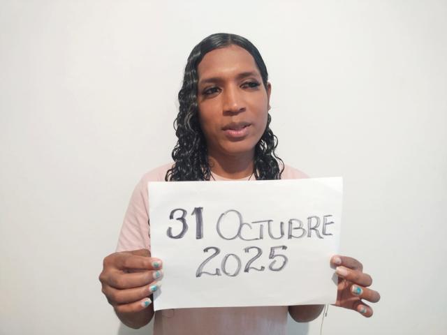 MiaCockTs with dark brown skin, black wavy hair, wearing a pink shirt, holding a sign reading "31 October 2025" in her hands, blue-painted nails.