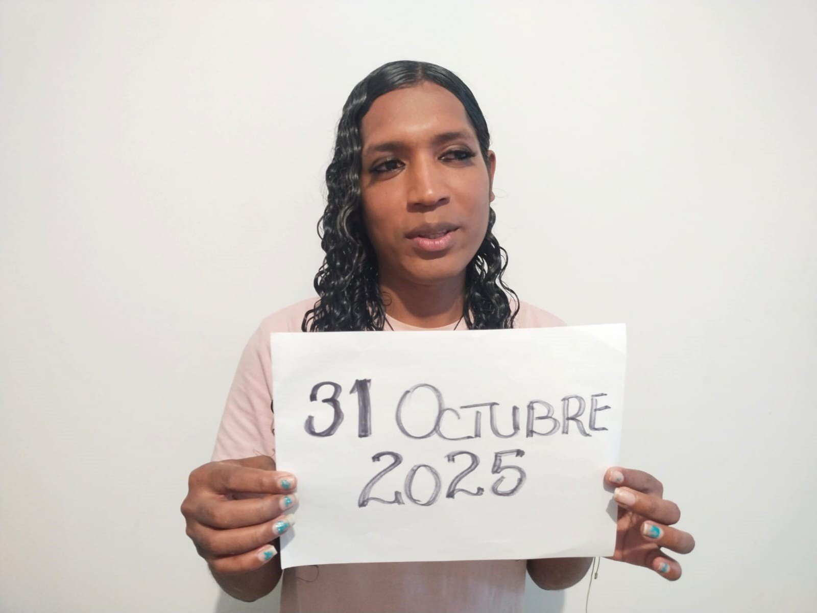 MiaCockTs with dark brown skin, black wavy hair, wearing a pink shirt, holding a sign reading "31 October 2025" in her hands, blue-painted nails.