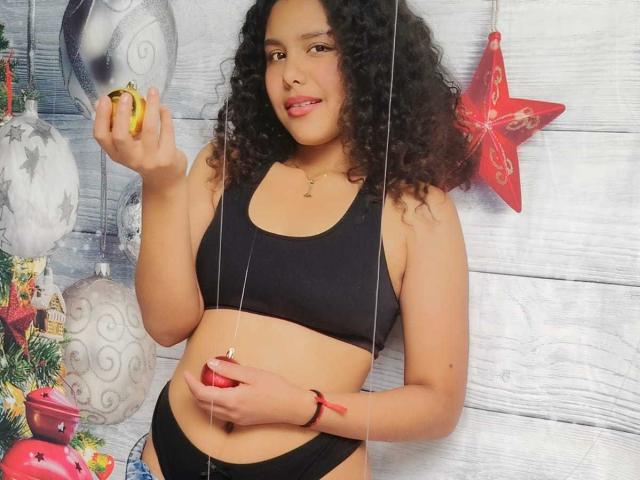 AriaVega with curly black hair, tan skin, medium tits in black sports bra and underwear, holding banana, red star ornament, white wood background, smiling, holding string.