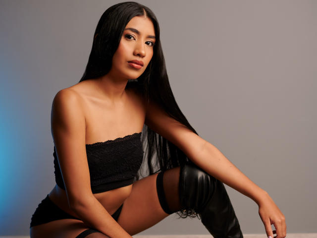 HottieEmily with long black hair, tan skin, and small tits, wearing a black lace tube top and thigh-high black boots, sitting with a relaxed, confident expression.