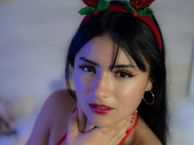 LizzeJoy with black hair, red lips, hoop earrings, and sparkly reindeer headband, blue-tinged face, red halter, hand on neck, looking down, medium skin, makeup, soft light.