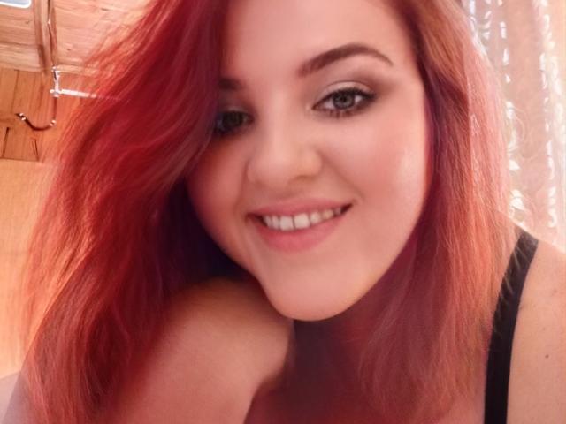 NatashaNikanorovna with red hair, smiling, wearing black straps, showing her shoulder and upper chest, brown eyes, and a cute nose. Her tits are partially visible.