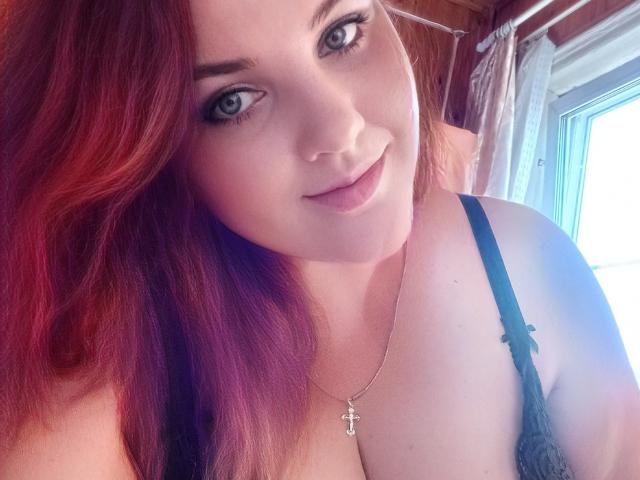 NatashaNikanorovna with red-purple hair, blue eyes, and black lace bra, smiling softly, wearing a silver necklace with a small cross, showing her tits.