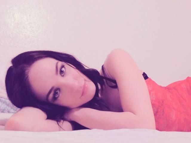 PhoenixPaulline lies on a bed, her black hair and blue eyes contrasting with a red lace dress, her bare shoulders and arm resting on white pillows.