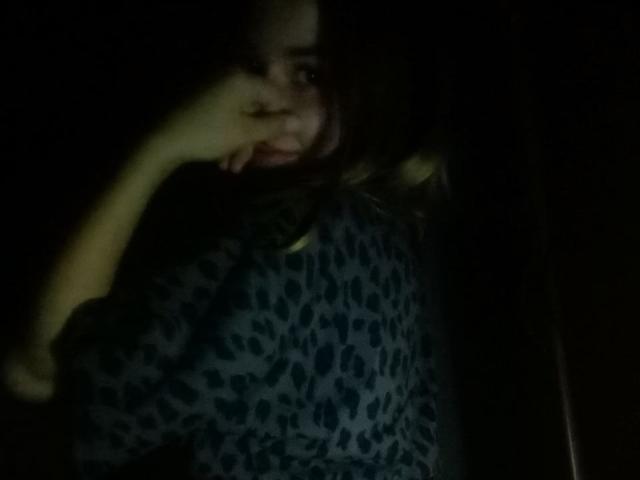 AileenShelly with dark hair, wearing a blue leopard print shirt, partially covering her mouth with her hand, in a dimly lit, shadowy setting.