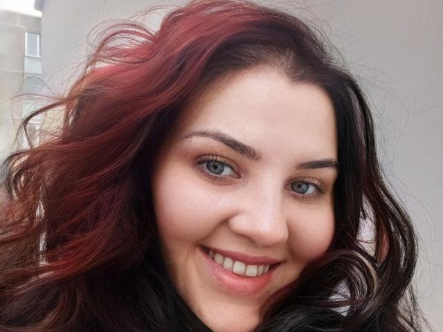 NatashaNikanorovna has wavy dark brown hair with red highlights, blue eyes, and a cute smile showing white teeth. She's wearing a black top. Her face is centered.