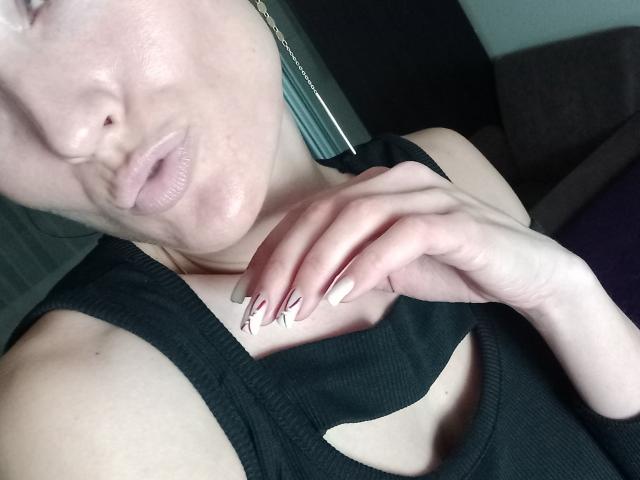 BellaJulls with puffy lips, wearing a black cut-out top, has her hand near her chin, showing off her French-manicured nails, pale skin, and subtle makeup.