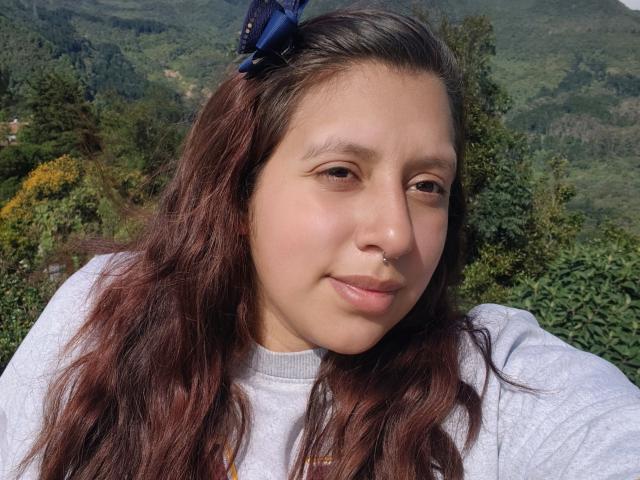 ArenaPeeach tiene el pelo castaño y ondulado, un clip azul para el pelo, un piercing en la nariz y una camiseta gris. Está sonriendo con una ligera inclinación de la cabeza. Su vulva no es visible..
