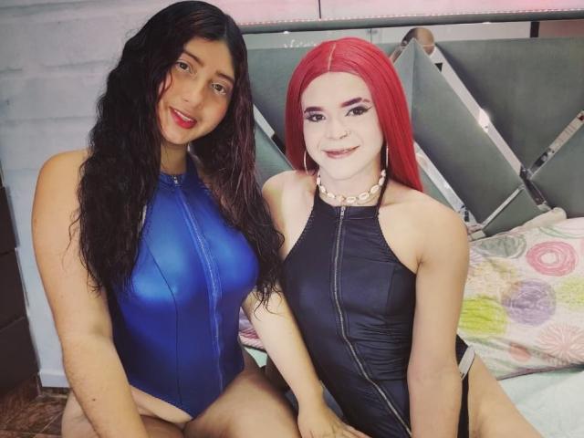 Two DiamondSexyy's sitting on a bed, both in shiny one-piece swimsuits. One has black hair and blue swimsuit, showing tits. The other has red hair and black swimsuit.