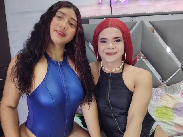 Two DiamondSexyy's with long hair, one with dark brown hair in a blue shiny leotard, and the other with red hair in a black shiny leotard, sitting together.