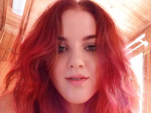 NatashaNikanorovna has red, wavy hair, pale skin, green eyes, and pink lips, looking at the camera with a slight smile. She's wearing a wooden roof in the background.