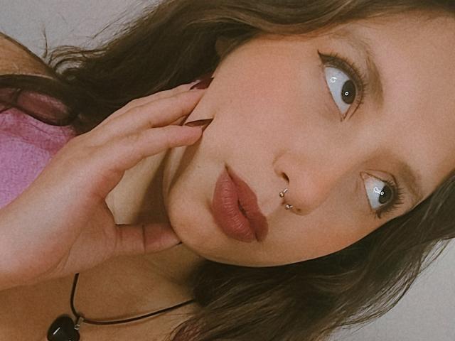ArenaPeeach with nose piercings, full lips, blue eyes, and long brown hair, touching her face with manicured nails, wearing a pink top, looking up with a slight smile.