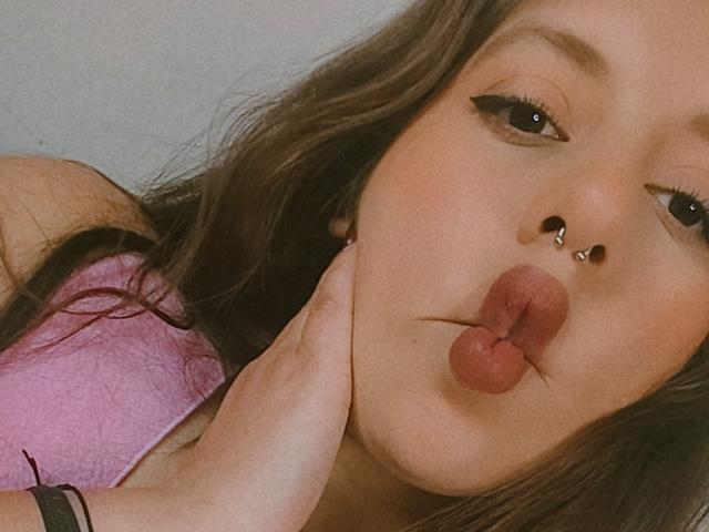ArenaPeeach heeft volle, bruine lippen, een neusring en een septumring, met haar hand op haar wang, draagt een roze top en heeft lang bruin haar..