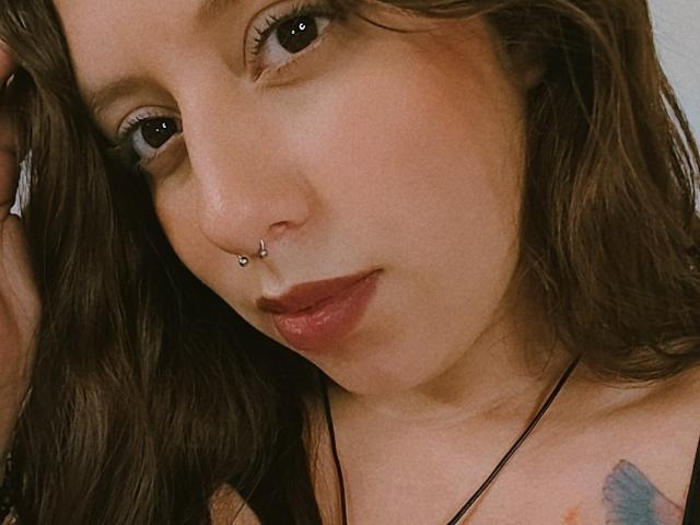 ArenaPeeach has a nose piercing with two small silver balls, brown wavy hair, and a tattoo on her right shoulder, with a black necklace and dark lipstick.