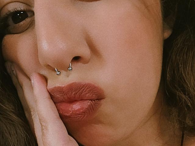 O rosto de ArenaPeeach, com um piercing no nariz, dois piercings nas narinas e lábios volumosos, avermelhados e marrons. Ela está tocando sua bochecha com um dedo, e seus cabelos castanhos ondulados aparecem ao fundo..