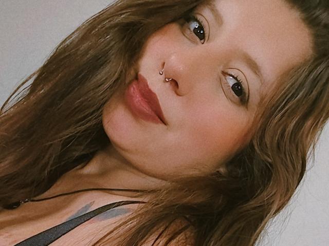ArenaPeeach heeft golvend bruin haar, een neusring en rode lippenstift, met een subtiele tatoeage op haar rechter schouder, en draagt een zwarte top met bandjes. Haar ogen zijn iets samengeknepen..