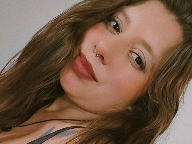 ArenaPeeach hat welliges, braunes Haar, eine Nasenpiercing und roten Lippenstift, mit dezentem Augen-Make-up. Ihre Lippen sind leicht geöffnet, wodurch ein Hauch ihrer Zähne sichtbar ist..
