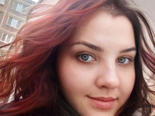NatashaNikanorovna's face with red and black wavy hair, blue eyes, and pink lips, showing a slight smile. Her nose is small, and her eyebrows are thick.