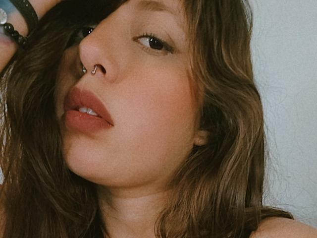 ArenaPeeach's face, showing brown wavy hair, nose ring, full lips, and slight smile. She's wearing a black bracelet on her right wrist. Soft light highlights her skin.