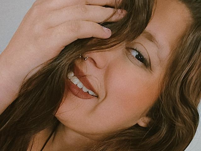 Il volto sorridente di ArenaPeeach, con i suoi capelli castani ondulati, il piercing al naso e i denti bianchi, è ben visibile. Sta toccando i capelli con la mano, mostrando un'espressione carina e giocosa..