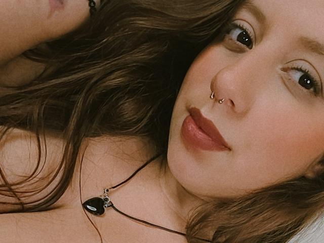 ArenaPeach tiene el cabello castaño y ondulado, un piercing en la nariz y lápiz labial oscuro, lleva un collar con un corazón negro, y está mirando a la cámara con una ligera sonrisa..